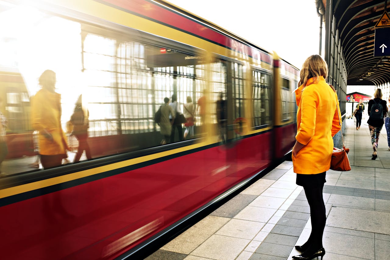 A woman in a yellow coat waits as a red train passes in a bustling station.